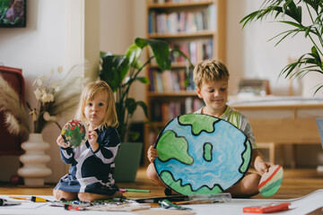 Siblings painting at home with watercolors and tempera paints, creating a model of planet Earth.