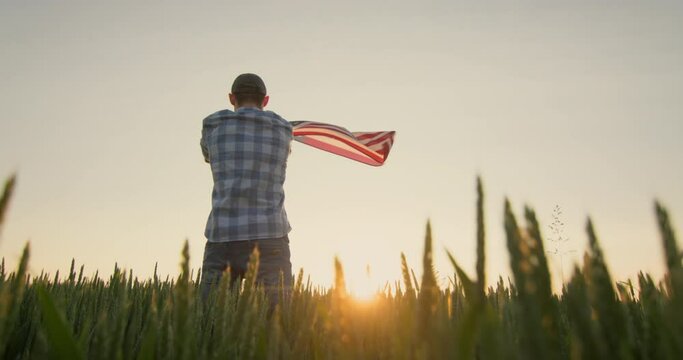 American farmer waving the US flag against the backdrop of a field of wheat where the sun rises. Low angle shot