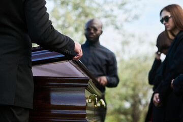 Close-up of man in black suit closing lid of coffin at funeral ceremony after farewell and saying...