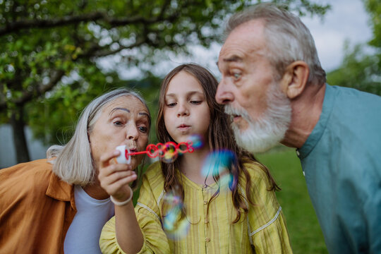 Grandparents and granddaughter are blowing bubbles from a bubble wand.
