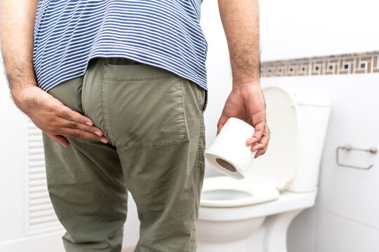 Male suffers from diarrhea holds toilet paper roll in front toilet bowl. Man squeezing tissue with diarrhea excreting in toilet. Hand holding a roll toilet paper is feeling pain with constipation.