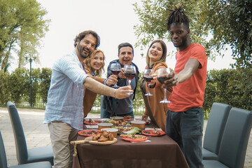 Multiethnic friends in a garden courtyard, raising wine glasses to toast directly with the viewer. Standing and smiling, they share the warmth of the evening.