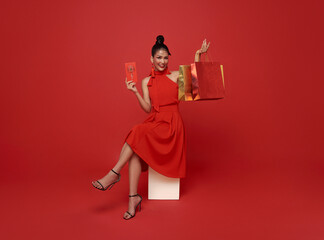 Happy Asian shopper woman wearing red dress sitting on chair holding shopping bag and angpao or red packet monetary gift isolated on red background. Shopping bless Chinese new year