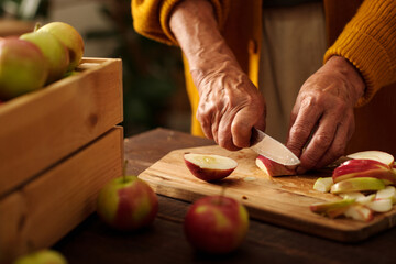 Hands of aged woman cutting fresh ripe apples with knife on wooden board while standing by table...