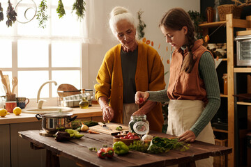 Cute girl in apron putting fresh tomato into open jar with marinade mix while helping her...