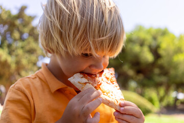 little blond boy eats pizza in the park sitting on the grass