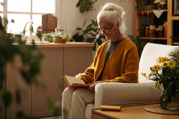Serious senior woman in woolen knitted yellow cardigan reading book while sitting in big...