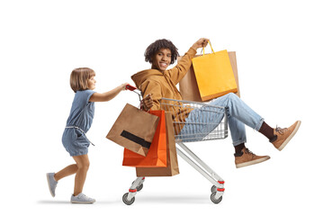 Little caucasian girl pushing an african american young man with shoppin bags inside a shopping cart