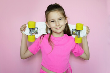 Happy girl is holding bright colored skateboard in her hands, clutching it to her and smiling playfully on bright pink background in isolation. Childhood and happy time