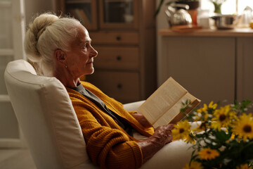 Serious grandmother with open book waiting for someone while sitting in comfortable armchair by bunch of yellow garden flowers in country house