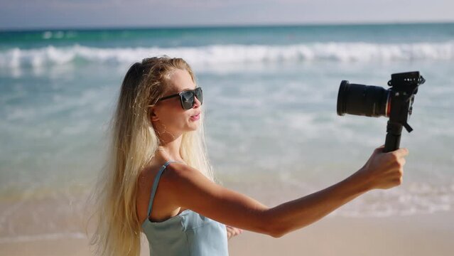 Woman travel vlogger adores summer vacation in Sri Lanka at ocean beach filming herself on camera. Young blonde blogger in sunglasses walking on sand holding professional movie camera. Slow motion.