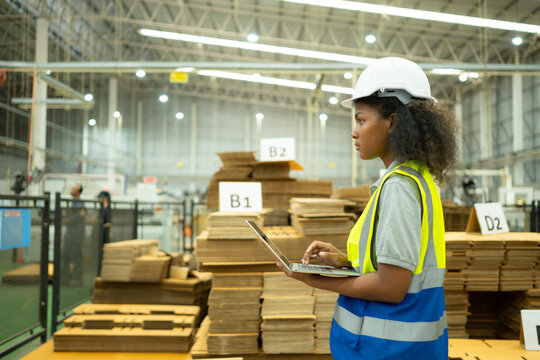 A young female warehouse worker checks and counting cardboard boxes in the warehouse using a laptop.