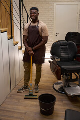 Smiling guy in a work apron cleans in a barbershop
