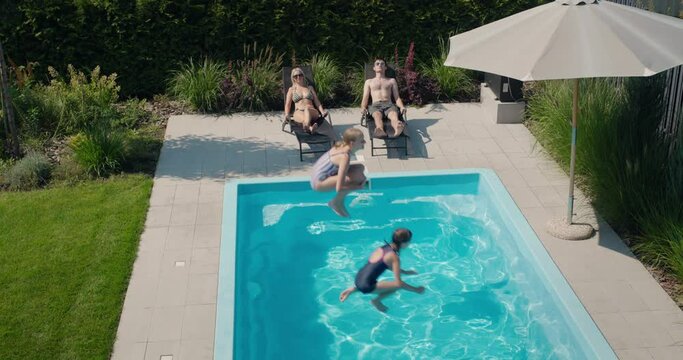 Children jump into the pool, slow motion video. Adults rest on sun loungers nearby.