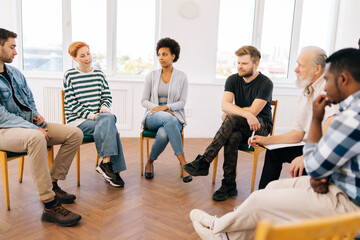 Front view of young woman telling about mental problem or addiction to other patients sitting in circle during group therapy session. Concept of mental health, psychotherapy, depression, social issues - obrazy, fototapety, plakaty