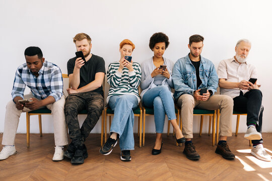 Front view of multicultural and different ages professional business people for vacancy in casual clothes sitting on chairs in row using mobile phones, while waiting job interview on white background