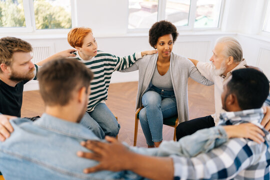 High-angle view of multicultural and different ages group of addicted people discuss problems and together holding shoulders each other on therapy sitting on chairs in circle. Concept of mental health