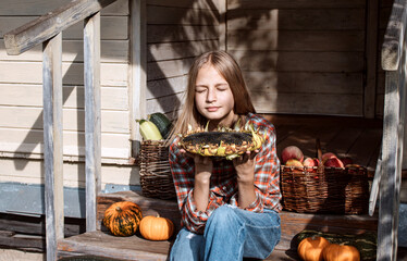 Girl is sitting on steps of an old village house holding large sunflower in her hands. Ripe pumpkins lie nearby and there are baskets filled with harvested crops: apples and zucchini. Harvest. Autumn.