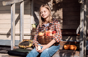 Girl is sitting on steps of an old village house with her face exposed to warm sunbeams holding the basket with apples. Ripe pumpkins, baskets with zucchini and big sunflower are nearby. Harvest. Fall