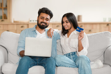Obraz premium E-commerce concept. Happy young indian spouses with laptop and credit card shopping online from home, sitting on couch