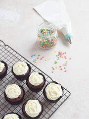 Homemade chocolate cupcakes with white buttercream on cooling rack. Piping bag with star-shaped nozzle and colored edible confetti for decorating pastries.