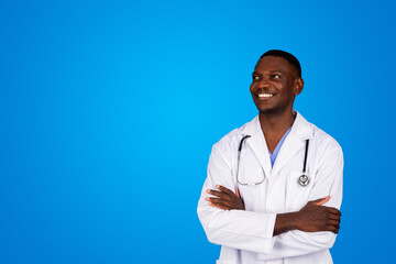 Smiling young african american guy doctor in white coat with crossed arms on chest looks at copy space