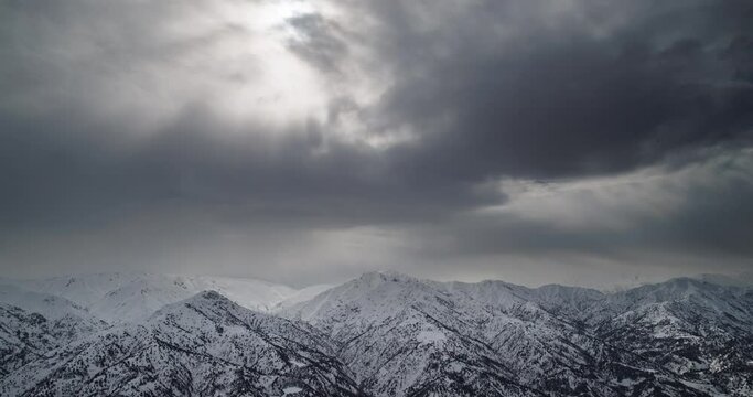 Winter mountains in snow timelapse. Snow-capped mountain ranges on cloudy day. Whole sky is in gray clouds rays of sun are barely visible behind clouds. In some places clouds part and can see blue sky