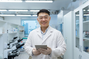 Portrait of a young Asian medical student. a scientist, a chemist who stands in the waiting room in a white coat and with a tablet in his hands, smiles at the camera.