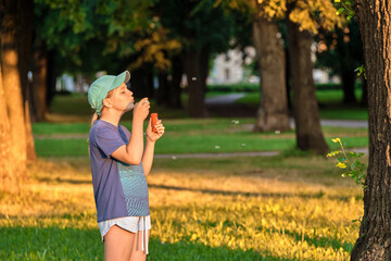 A girl of 10-12 years old blows soap bubbles in the park at sunset. School-age child enthusiastic playing outdoors.