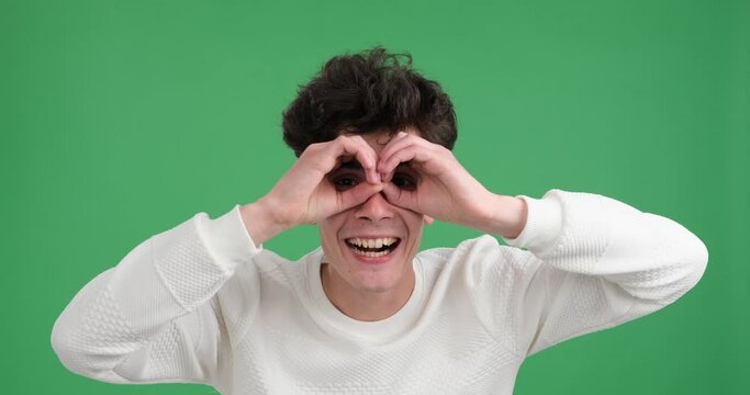 Caucasian man as he playfully gazes through an imaginary binoculars against a vibrant green backdrop. With a wide smile on his face and an enthusiastic posture, he embraces the spirit of curiosity.