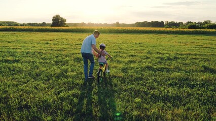 Careful father supports little daughter learning to ride bicycle in sunset field