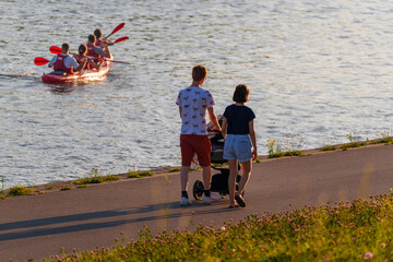 A happy family with a baby stroller walks along the embankment by the river at sunset. Modern...