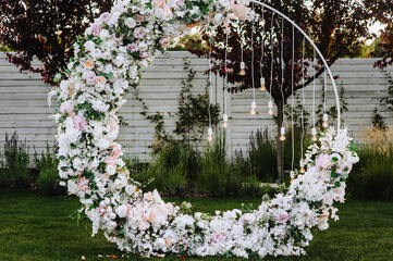 A beautiful wedding arch decorated with flowers, electric lamps, garlands stands in the garden outdoors in the evening.
