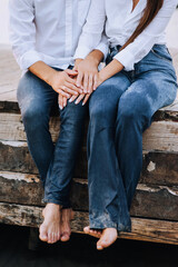 Barefoot man and woman in white shirts, blue wet jeans sitting on a wooden pier near the sea in summer. Close-up photography, portrait, vacation lifestyle.