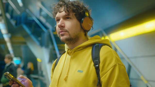 Young curly-haired man with backpack listening to music with headphones, moving his head to rhythm and using app on smartphone while waiting for a train on subway platform