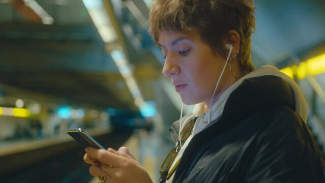 Young woman in earphones standing on subway platform, waiting for a train and texting on a smartphone. Side view, chest-up shot