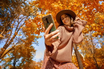 Autumnal style trendy shot. Stylish woman in hat and knit sweater captures the moment with selfie on smartphone at park with orange leaves