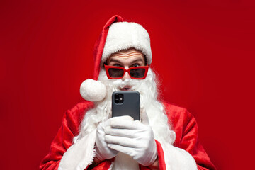shocked santa claus in hat and festive glasses uses smartphone and is surprised on red background