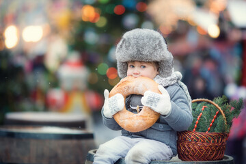 Little boy in grey winter clothes on Christmas fair eating huge bagel