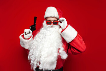 santa claus in hat and festive glasses holds gun in his hands on red background, dangerous man in santa costume