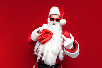 rude santa claus in cap and festive glasses showing middle finger on red background, angry man in...