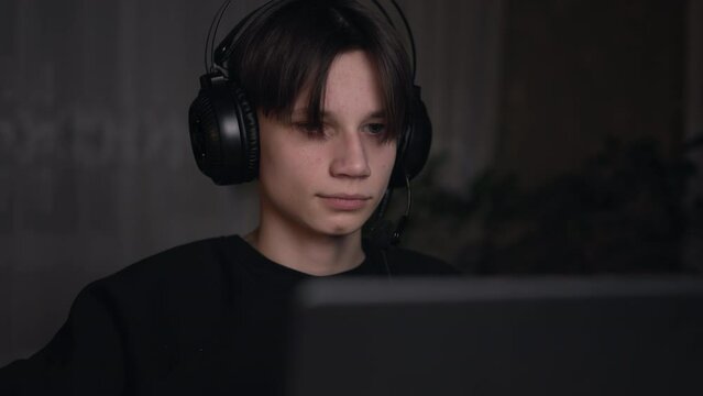 Front view of young computer gamer wearing headphones playing pc games, communicating in live stream in a dark room. Generation z, teen boy using laptop for esports or virtual sports