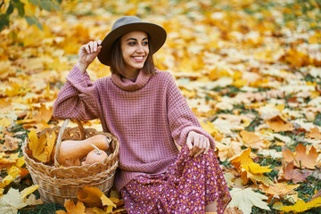 Stylish woman in cozy knitted sweater, elegant dress, and chic hat sits beside a basket with pumpkins. Surrounded by golden autumn leaves on the ground