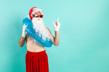 Fototapeta na wymiar santa claus in suit with inflatable swim ring in sunglasses rests on the beach and points to the copy space