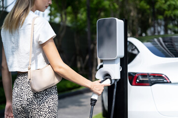 Young woman travel with EV electric car charging in green sustainable city outdoor garden in...