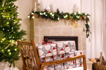 Merry Christmas! Stylish wrapped christmas gifts on arm chair against christmas tree with festive lights and cozy burning fireplace. Atmospheric Christmas eve, holiday time.