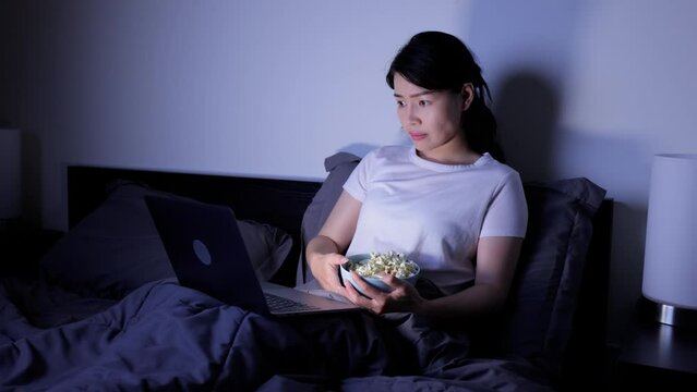 Asian woman eating popcorn while watching a scary movie on her laptop while sitting in bed.