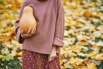 Faceless woman in sweater holding rape pumpkins surrounded orange leaves, enjoying autumnal time and seasonal harvest