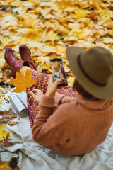 Back view of stylish woman in hat taking photo on camera phone while relaxing in beautiful autumn park