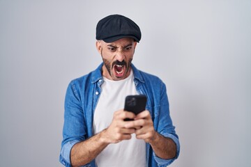 Hispanic man with beard using smartphone typing message angry and mad screaming frustrated and...